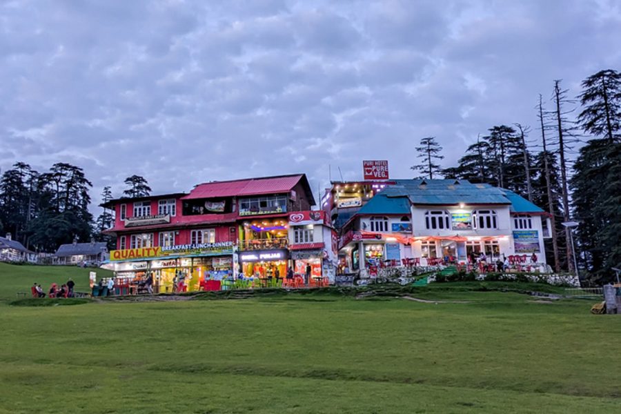 Dalhousie Tour Package, A vibrant evening view of a row of colorful buildings with illuminated signage in Dalhousie Khajjiar, known as Mini Switzerland, against a backdrop of tall pine trees and a twilight sky.